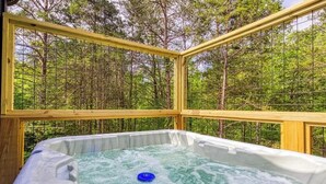 Un jacuzzi con un objeto flotante azul, rodeado por una terraza de madera y una cerca de malla metálica, con un bosque de fondo.