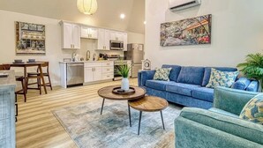 Una sala de estar moderna con un sofá azul, una mesa de comedor con sillas, una zona de cocina con armarios y electrodomésticos blancos, y un cuadro grande en la pared.