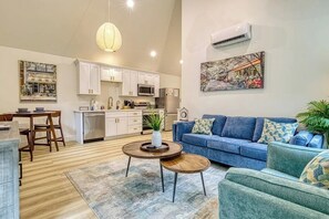 A modern living room with a blue sofa, a dining table with chairs, a kitchen area with white cabinets and appliances, and a large painting on the wall. - Wooded Couples Retreat • Private Hot Tub (Sevierville)