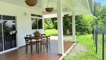 An outdoor patio with a wooden table and chairs, a hanging basket, and a view of a green garden.