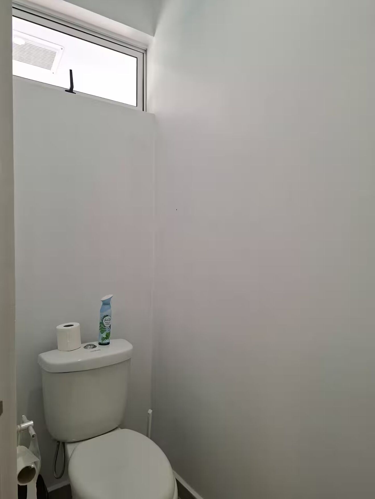 Un baño pequeño y blanco con inodoro, ventana y rollo de papel higiénico.