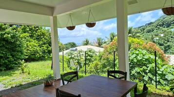A covered outdoor dining area with a table and chairs, surrounded by lush greenery and a mountainous backdrop.