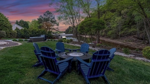 A patio with Adirondack chairs and a round table, surrounded by a grassy lawn and trees. - Belle Pines Hideaway by Avantstay Lakefront + Hot Tub + Fire Pit + Game Room Near Charlotte (Sherrills Ford)