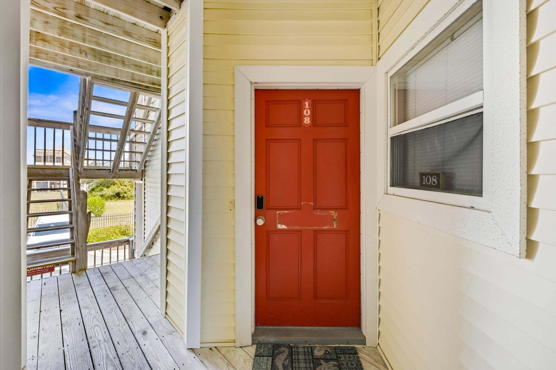 A red door with a number plate, a wooden porch, and a window with a number plate.