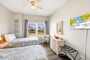 A bedroom with two beds, a ceiling fan, a window with a view of houses, a TV on the wall, and a wicker dresser. - Rogue KDH steps to the  Beach condo w/ pool (Kill Devil Hills)