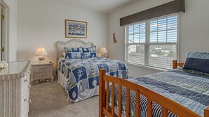 Quarto com uma grande janela, cama com colcha azul, mesa de cabeceira com abajur e cadeira de vime.