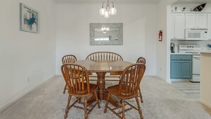 Uma sala de jantar com mesa e cadeiras de madeira, um candelabro, um espelho e uma cozinha ao fundo.