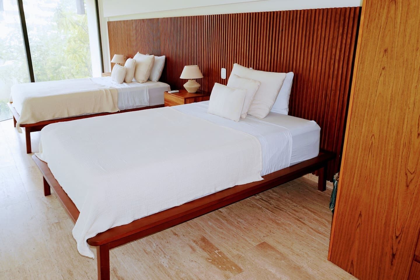 A hotel room with two beds, wooden headboard, white linens, and a nightstand with a lamp.