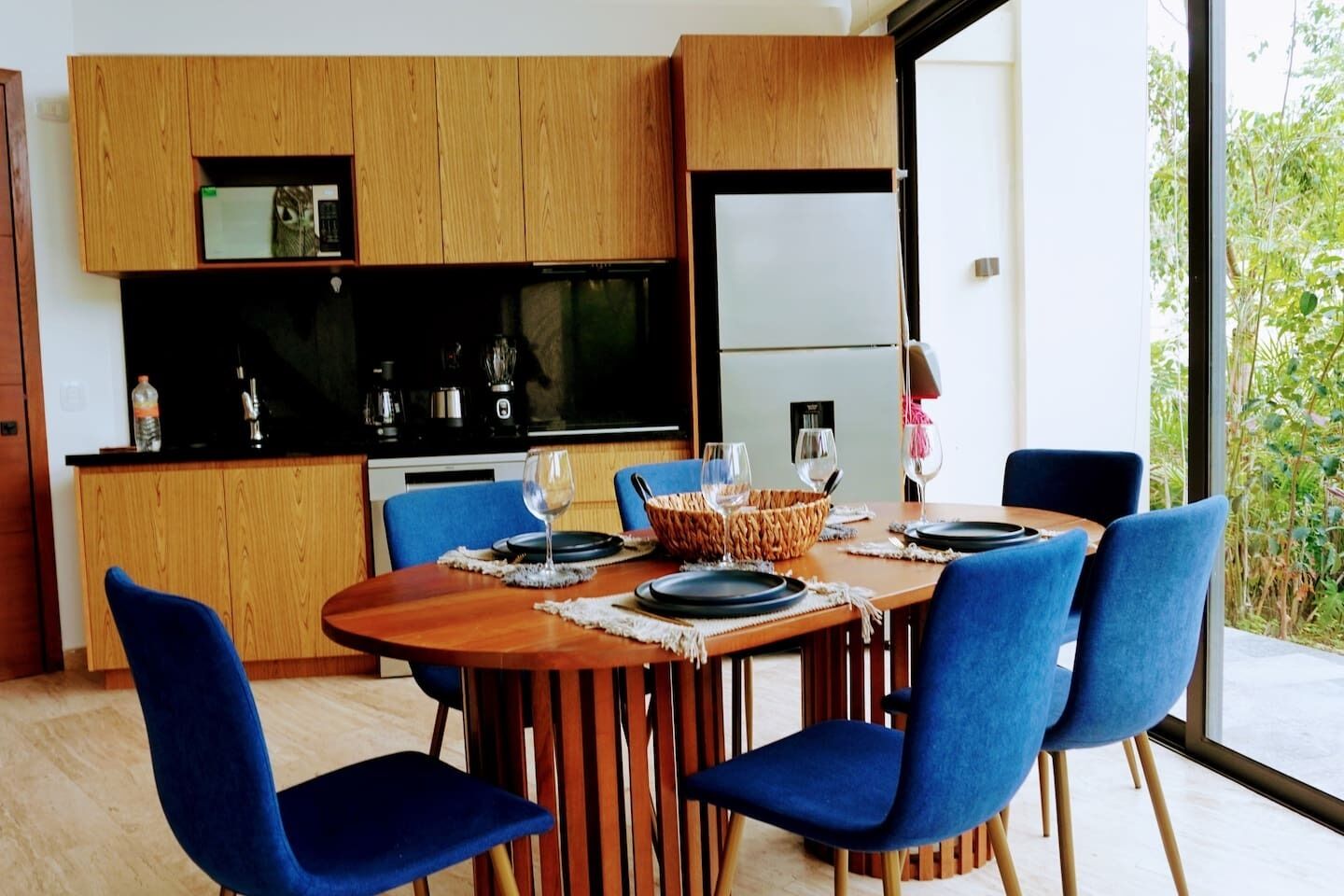 A modern kitchen with wooden cabinets, a dining table with blue chairs, and a view of greenery outside.