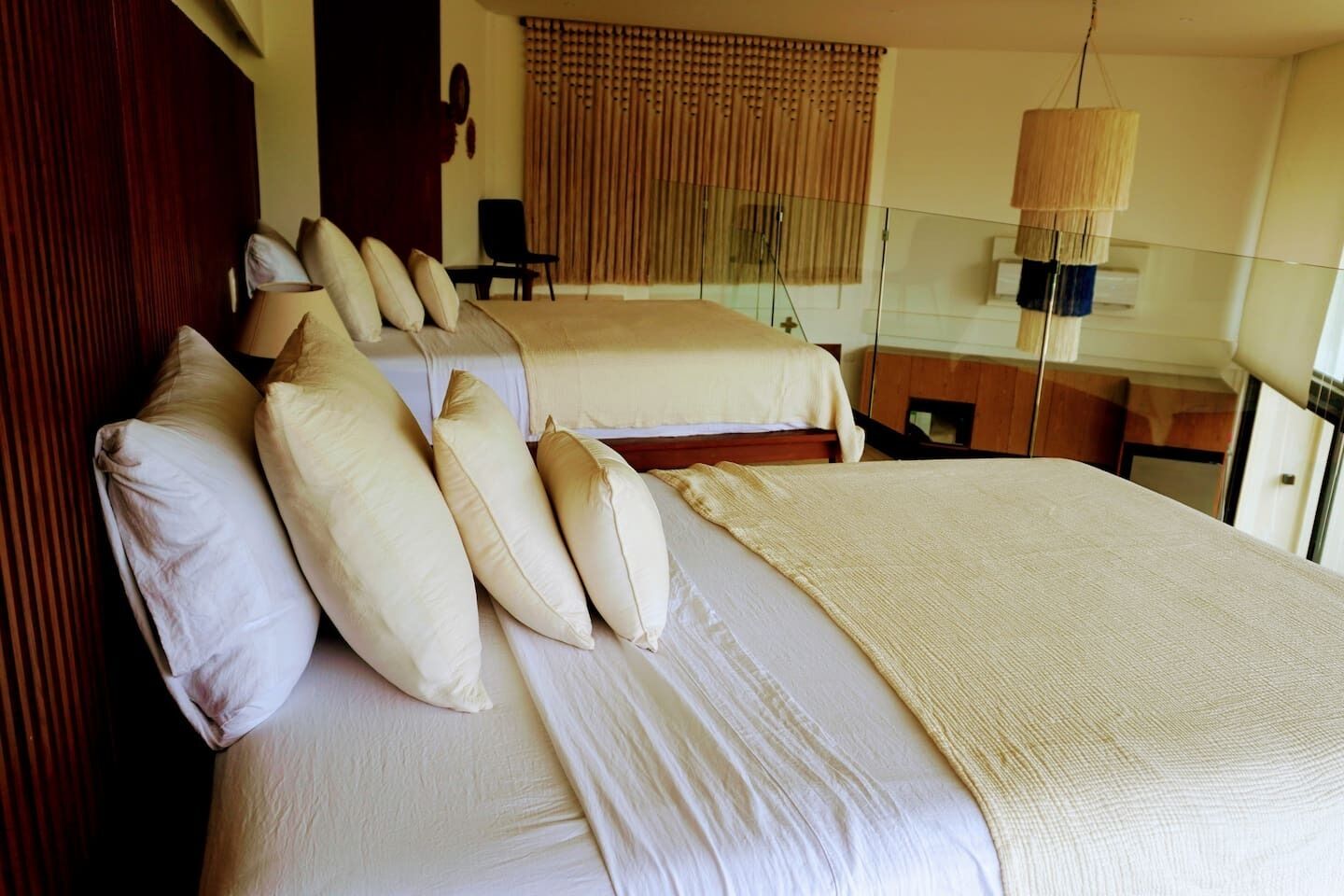 A hotel room with two beds, a wooden headboard, a large window with blinds, a hanging lamp, and a desk with a chair.