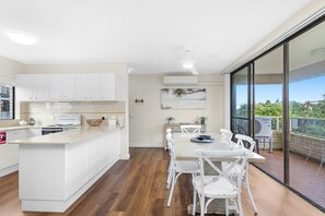 A modern kitchen with white cabinetry, a dining area with a large table and white chairs, and a view of a balcony with a view of the outdoors. - Sunset Reset Beachfront in Soldiers Point (Soldiers Point)