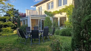 Pihalla on patio, jossa on pöytä ja tuoleja, katettu oleskelualue sekä rakennus, jossa on suuret ikkunat ja parveke.