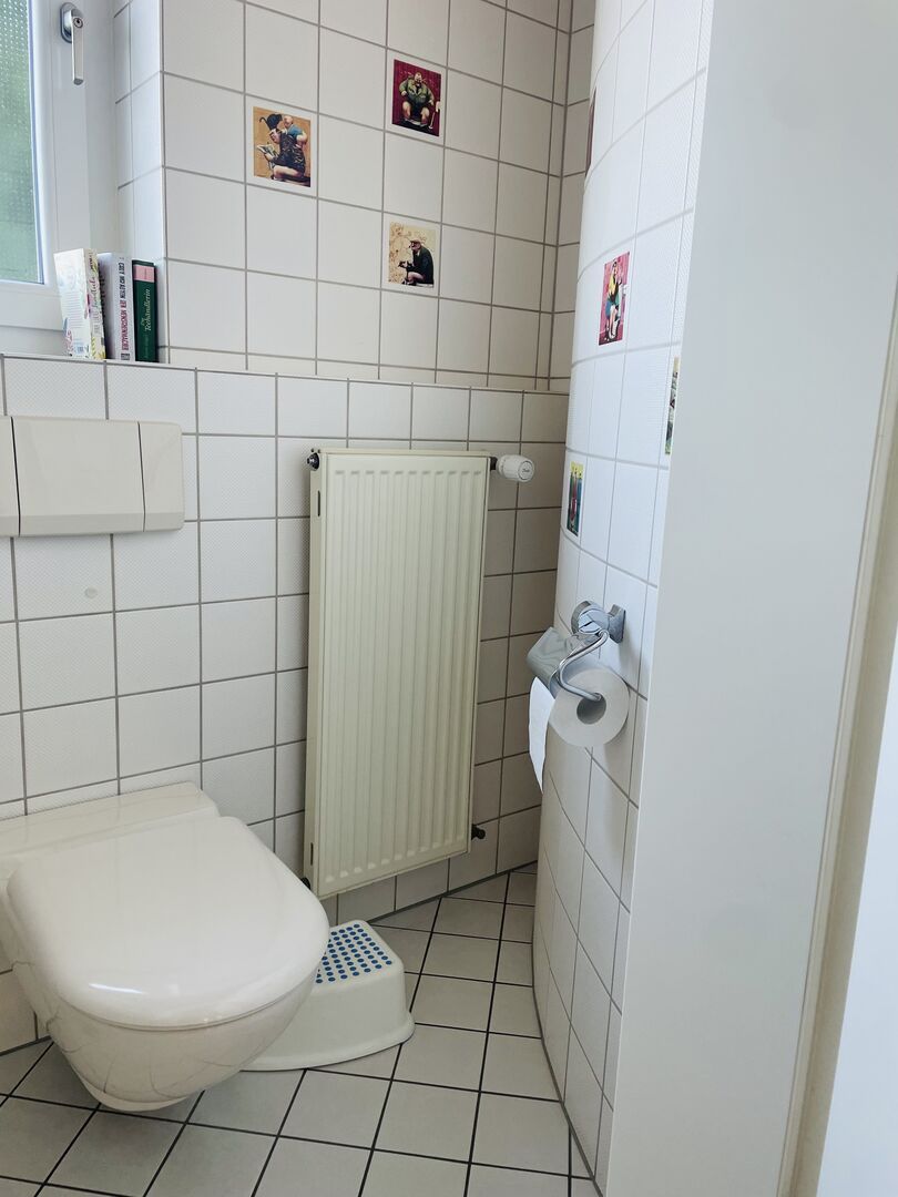 A small, white bathroom with a toilet, a radiator, and a shelf with books.