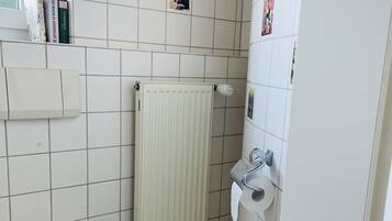 A small, white bathroom with a toilet, a radiator, and a shelf with books.