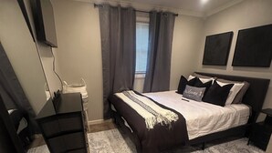 Quarto com cama, escrivaninha, cadeira, televisão e janela com cortinas.