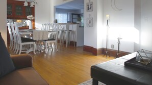 Een ruime woonkamer met een bruine bank, een eethoek met witte stoelen, en een keuken op de achtergrond.