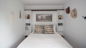 Een slaapkamer met een bed, een aan de muur hangend schilderij, planken met decoratieve objecten en twee hoeden aan de wand.