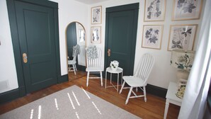 Een gang met twee groene deuren, witte stoelen, een klein tafeltje met een vaas en botanische prints in lijsten aan de muur.
