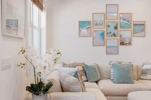 A living room with a beige sofa, a potted orchid, and framed beach-themed artwork on the wall.