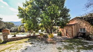 A stone building with a tiled roof, a gravel path, and a large tree in a garden setting. - Authentique maison piscine (Laroque Des Albères)