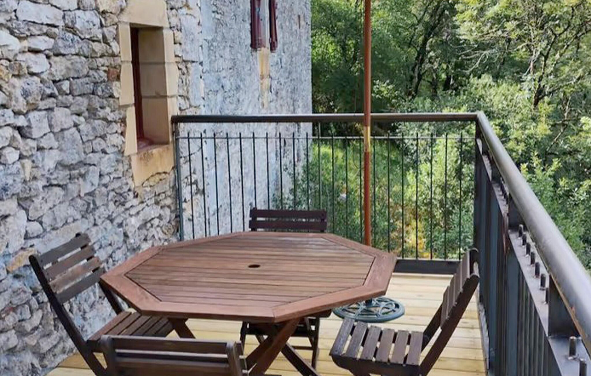 A wooden deck with a table and chairs, a stone wall, and a metal railing overlooking a forested area.