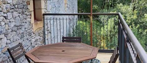 A wooden deck with a table and chairs, a stone wall, and a metal railing overlooking a forested area.