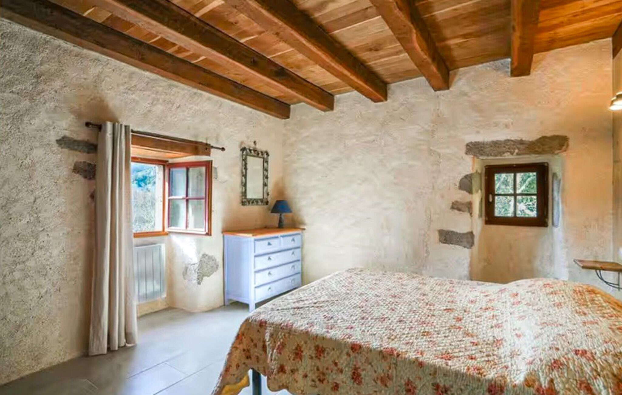 A rustic bedroom with a wooden ceiling, a bed with a floral bedspread, a dresser, a mirror, and a small window.
