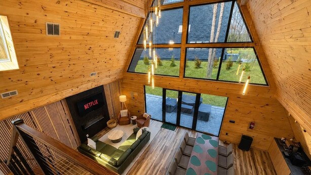 A wooden cabin interior with a fireplace, seating area, and large windows overlooking a garden. - A-Frame Escape | Firepit Nights + Lake Days (Coolbaugh Township)