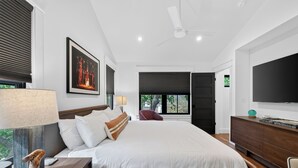 Un dormitorio moderno con una cama grande, un televisor de pantalla plana, un ventilador de techo y una ventana con vista a follaje.