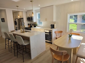 Una cocina moderna con una isla central, taburetes de bar, una mesa de comedor con sillas y vistas al exterior.