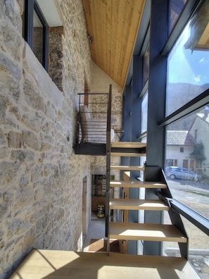 Un intérieur moderne avec un mur en pierre, un escalier en bois et de grandes fenêtres offrant une vue sur l’extérieur.