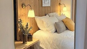 Makuuhuoneessa on puinen seinä, valkoisilla vuodevaatteilla varustettu sänky, kaksi yöpöytävalaisinta ja pieni kasvi yöpöydällä.