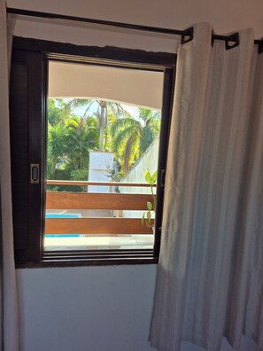 Ein Fenster mit Blick auf ein Schwimmbad, eine Holzterrasse und viel Grün.
