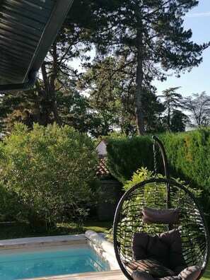 Un fauteuil suspendu au bord d’une piscine, avec vue sur un jardin et des arbres.