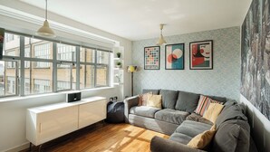A modern living room with a grey sofa, wooden flooring, a white radiator, and a large window with blinds. - Bright Spacious 1BR Flat in Vibrant Dalston (London)