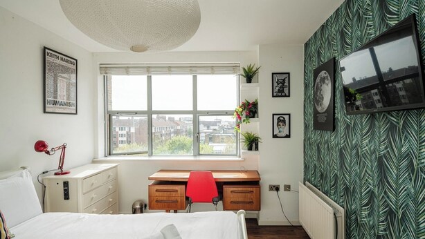 A bedroom with a bed, a desk, a chair, a window with a city view, and a wall with a leaf pattern. - Bright Spacious 1BR Flat in Vibrant Dalston (London)