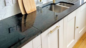 Una cocina con cubierta negra, armarios blancos, grifo dorado y tabla de cortar de madera.