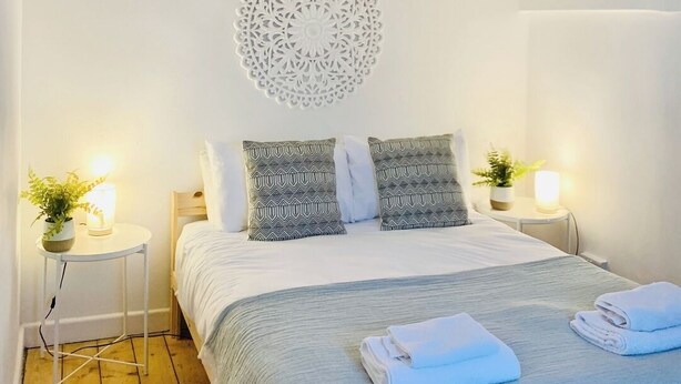 Un dormitorio con cama, almohadas, decoración en la pared, mesitas de noche y una planta.