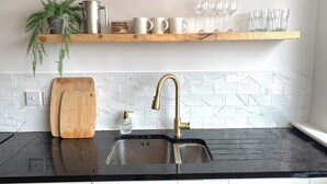 Una cocina con encimera negra, un lavamanos de acero inoxidable, una tabla de cortar de madera, una planta en maceta y una repisa con tazas y vasos.