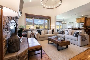A spacious living room with a stone fireplace, a large window with a mountain view, a kitchen with wooden cabinets, and a cozy seating area. - Perfect Summer Getaway! Recreation Meets Relaxation- Minutes to Lake! (Heber City)