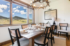 Un comedor con una mesa grande dispuesta para seis personas, sillas, una lámpara de araña y una ventana con vistas a un valle.