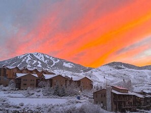 Une montagne enneigée, un ciel embrasé par le coucher de soleil, des bâtiments en bois aux toits couverts de neige et une vue dégagée sur la montagne.