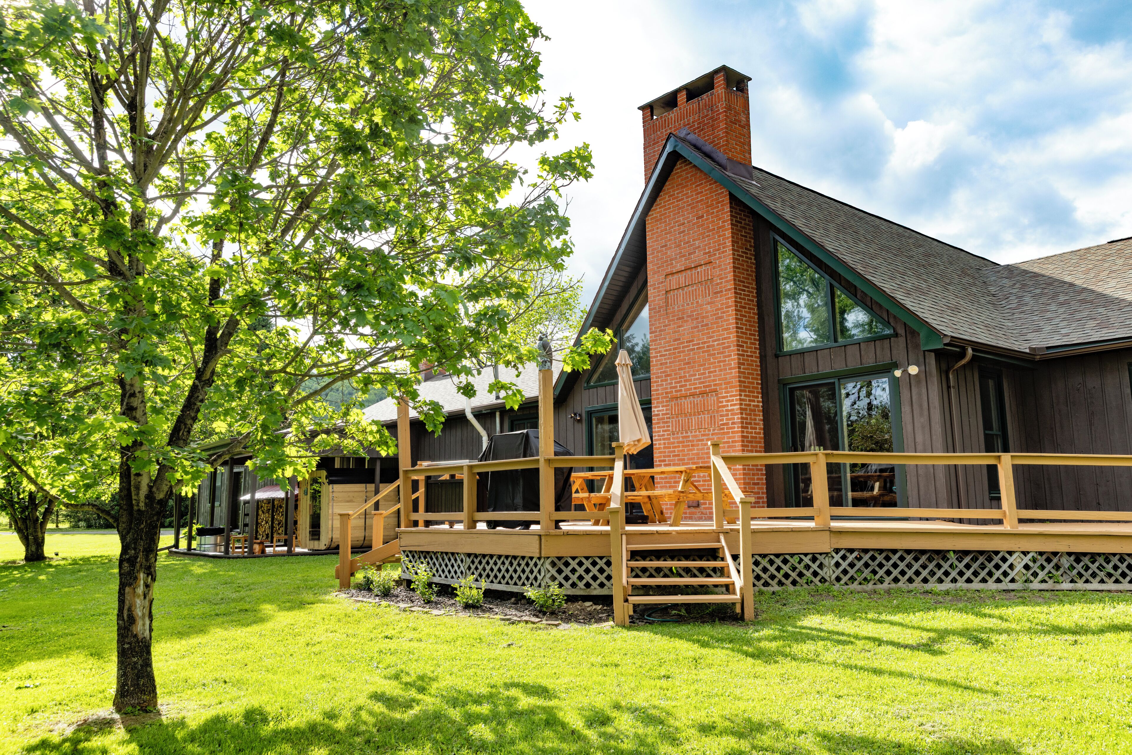 Un bâtiment avec une cheminée en briques, une terrasse en bois et un grand arbre au premier plan.