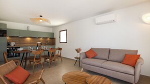 Een moderne woonkamer met een bank, twee houten bijzettafeltjes, rieten stoelen, een eettafel met stoelen, een keukenhoek en een plafondventilator.
