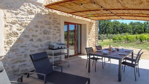 Een stenen gebouw met een patio, inclusief een tafel, stoelen, een barbecue en uitzicht op een wijngaard.