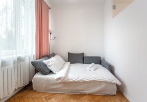 Un lit bien fait, avec des oreillers et une serviette enroulée, une fenêtre avec des rideaux légers et une lampe sur pied.