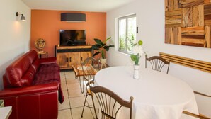 Un salon avec un canapé rouge, un téléviseur, un meuble en bois, une table à manger avec des chaises et une fenêtre ornée de plantes.