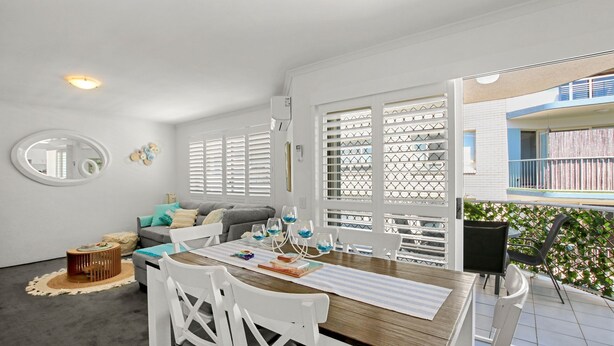 A dining area with a wooden table, white chairs, and a sofa in the background. - Ocean Breeze Unit 10, 42 King Street, Kings Beach (Kings Beach)