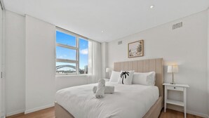 A hotel room with a large bed, a nightstand, a lamp, a framed picture on the wall, and a window with a view of a bridge. - KozyGuru | Iconic Panorama | Harbour Front Living (Lavender Bay)