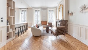 Ein geräumiges Wohnzimmer mit Fischgrätparkett, einem weißen Sofa, einem braunen Sessel und einem Kamin.